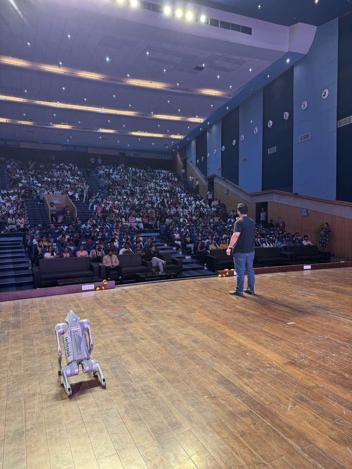 Gourav Gupta on stage in a large auditorium, with a robot on the stage floor beside him